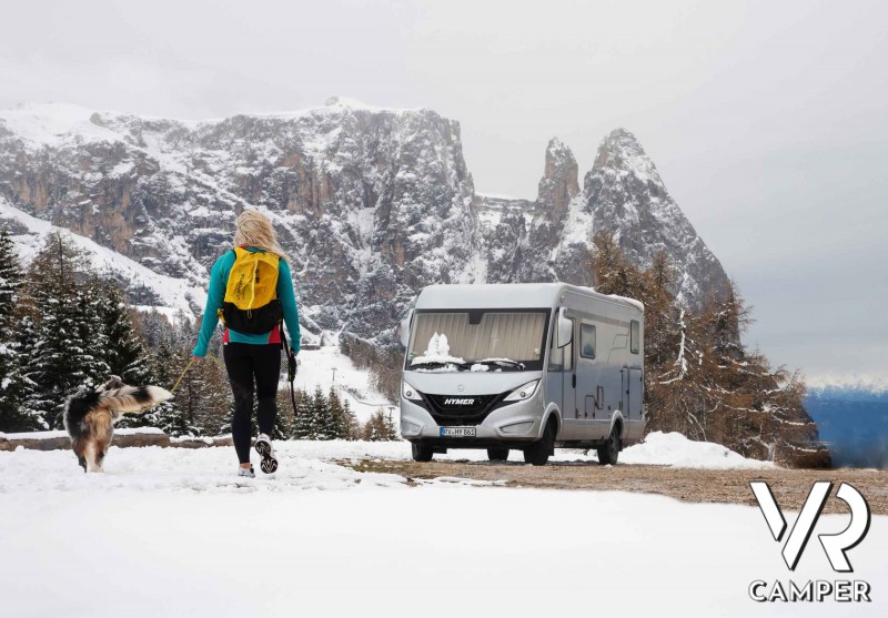 panorama con camper tra le montagne innevate e donna con zaino giallo e cane