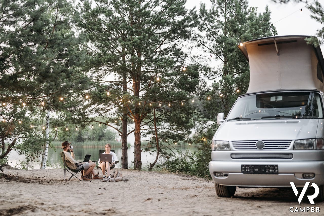Smart working in camper, furgonato parcheggiato nella natura e coppia che lavora al pc seduta fuori sul prato